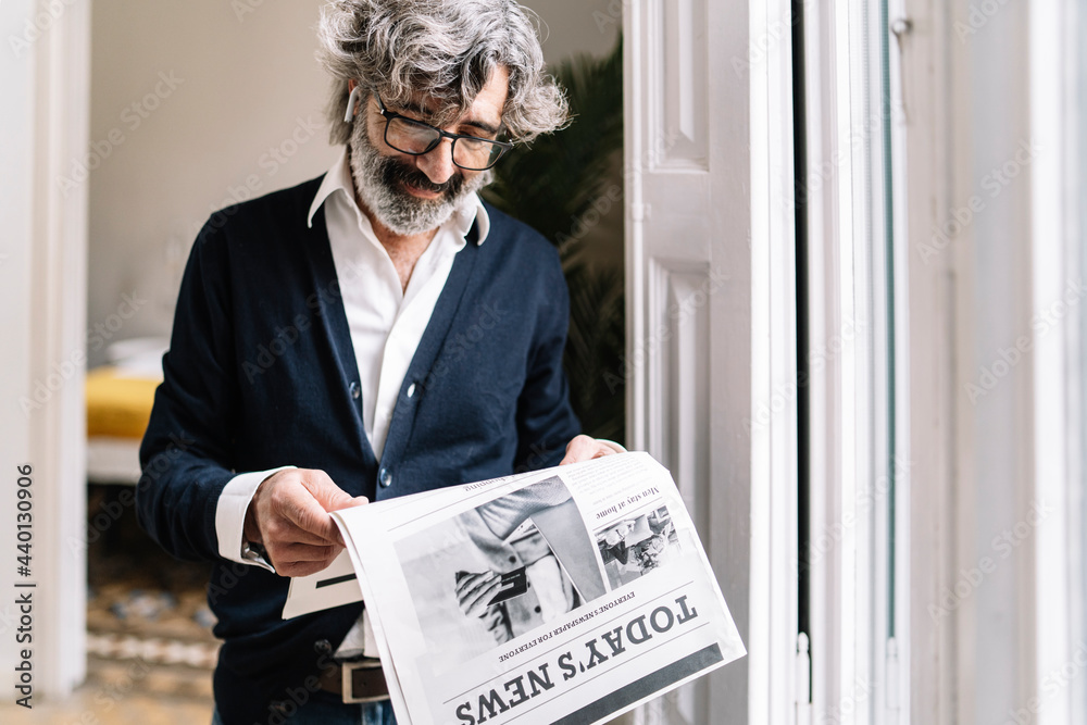 © COROIMAGE/Westend61 - Senior man with in-ear headphones reading newspaper at home