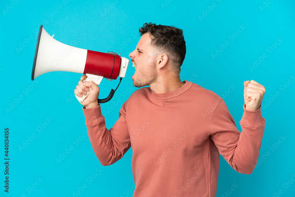 Young caucasian handsome man isolated on blue background shouting through a megaphone to announce something in lateral position Young caucasian handsome man isolated on blue background shouting through a megaphone to announce something in lateral position