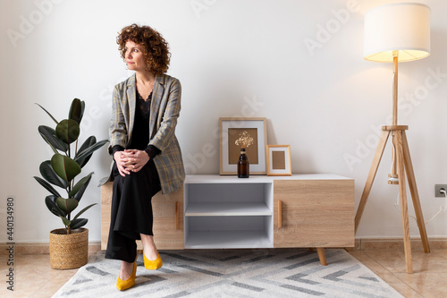 Mature woman looking away while sitting on wooden cabinet at home photo