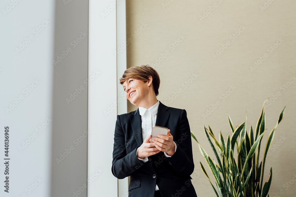 © Eugenio Marongiu/Westend61 - Happy female business professional looking away while holding mobile phone against beige wall in office