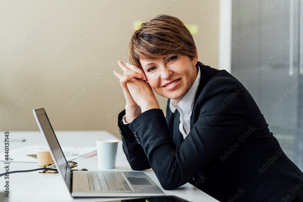 Happy businesswoman with hands clasped sitting in front of laptop at desk in office Happy businesswoman with hands clasped sitting in front of laptop at desk in office