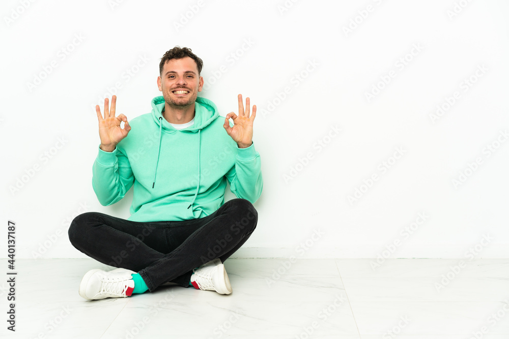 Fototapeta premium Young handsome caucasian man sitting on the floor showing an ok sign with fingers