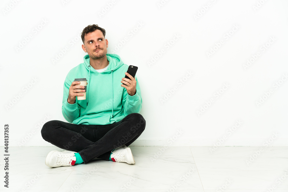 Young handsome caucasian man sitting on the floor holding coffee to take away and a mobile while thinking something Young handsome caucasian man sitting on the floor holding coffee to take away and a mobile while thinking something
