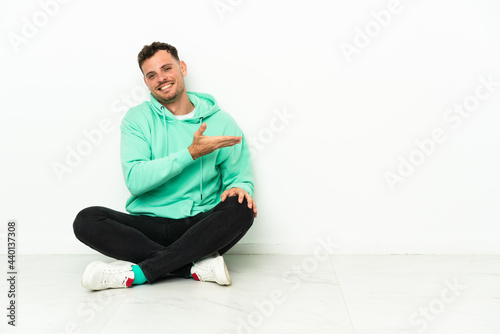 Young handsome caucasian man sitting on the floor presenting an idea while looking smiling towards