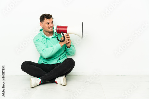 Young handsome caucasian man sitting on the floor shouting through a megaphone