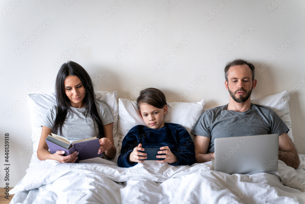 Woman reading book while father and son using wireless technologies in bed at home Woman reading book while father and son using wireless technologies in bed at home