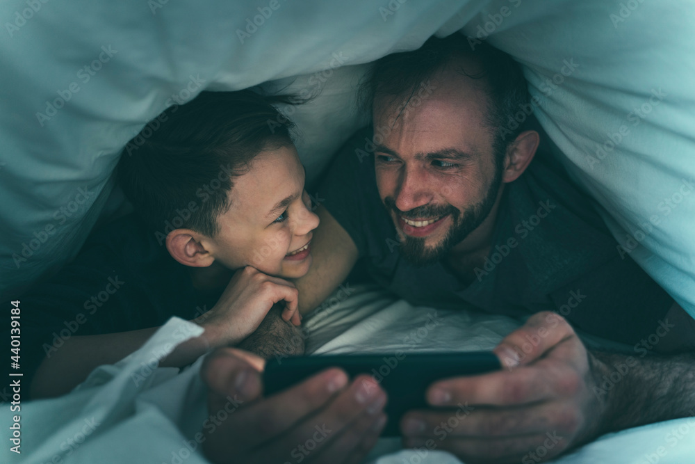 Father and son looking at each other while using smart phone under blanket at home Father and son looking at each other while using smart phone under blanket at home