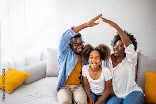 New building residential house purchase apartment concept. Stylish full family with kid sitting on sofa, mom and dad making roof figure with hands arms over head. Concept housing young family. photo