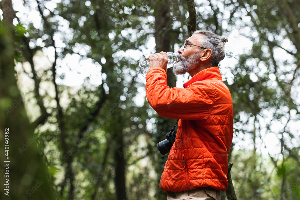Senior man with camera drinking water while standing in forest Senior man with camera drinking water while standing in forest