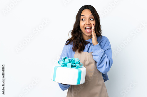 Pastry chef holding a big cake over isolated white background pointing to the side to present a product and whispering something