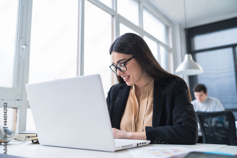 Female entrepreneur smiling while sitting in front of laptop with male colleague working in background at coworking office Female entrepreneur smiling while sitting in front of laptop with male colleague working in background at coworking office