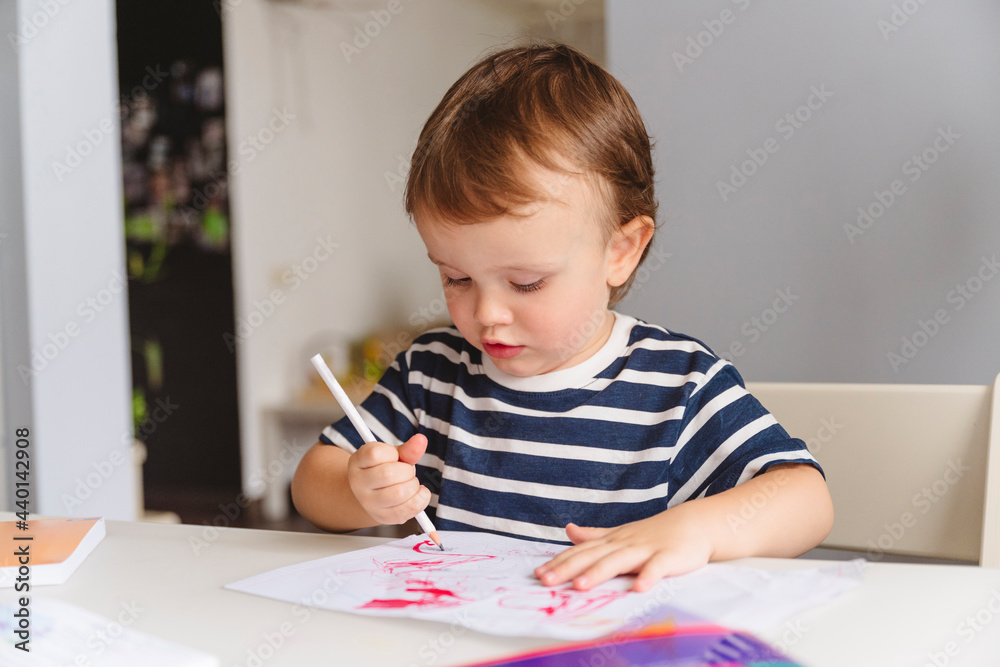 Cute boy drawing on paper with pencil sitting on table at home Cute boy drawing on paper with pencil sitting on table at home