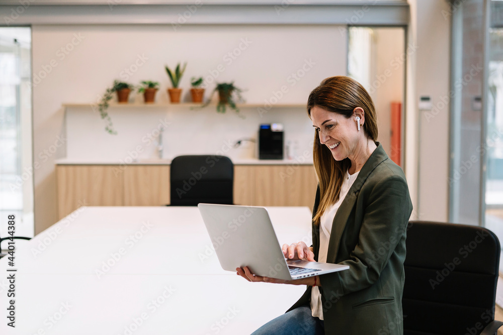 Smiling female entrepreneur with in-ear headphones using laptop while leaning on table at office Smiling female entrepreneur with in-ear headphones using laptop while leaning on table at office