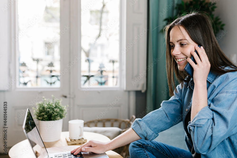 Smiling female entrepreneur talking on mobile phone while working on laptop at home office Smiling female entrepreneur talking on mobile phone while working on laptop at home office