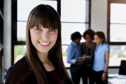 Smiling beautiful businesswoman with bangs standing while male and female colleagues discussing in office photo