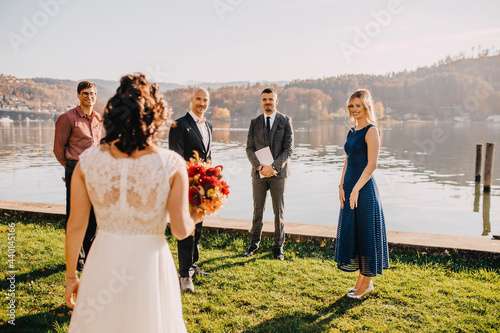 Groom standing with wedding guests looking at bride in front of lake on wedding ceremony photo