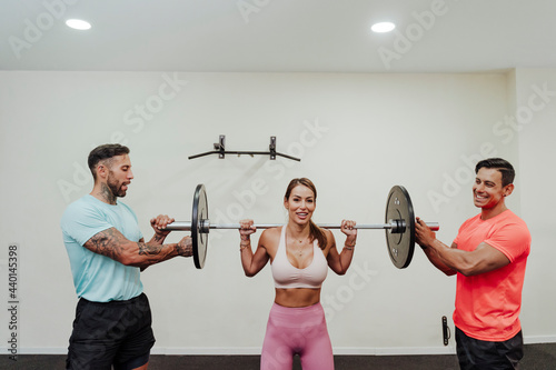Smiling female athlete lifting barbell with help of friends in gym photo