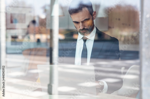 Serious businessman using laptop seen through glass window at office