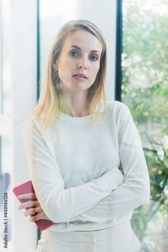 Mid adult confident businesswoman standing with arms crossed by window in office Mid adult confident businesswoman standing with arms crossed by window in office