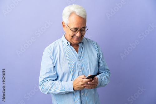 Middle age Brazilian man isolated on purple background sending a message with the mobile