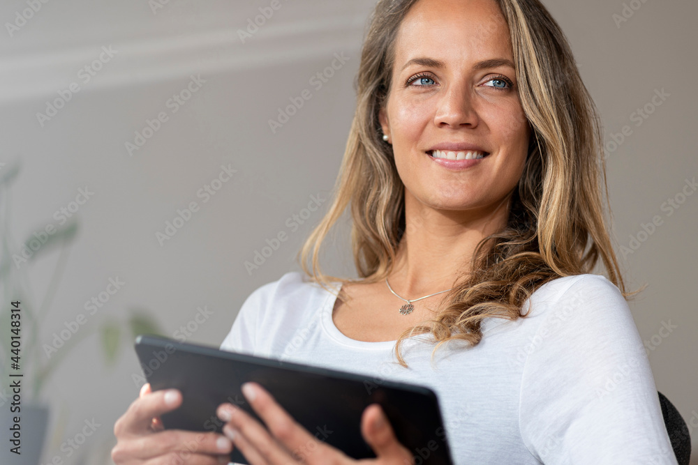 Smiling woman with blue eyes looking away while holding digital tablet at home Smiling woman with blue eyes looking away while holding digital tablet at home