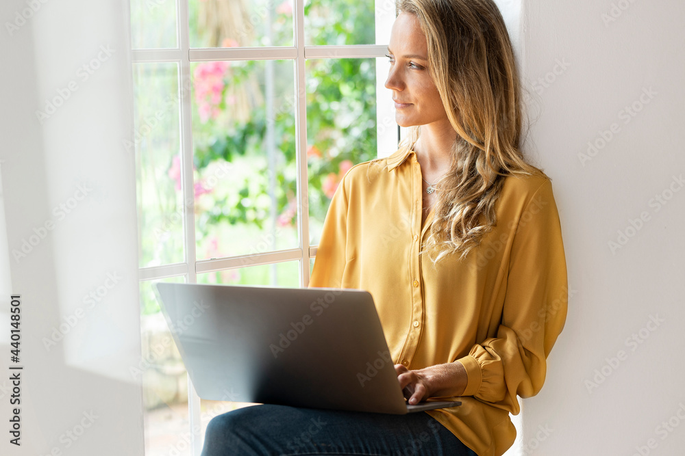 Contemplating woman looking away while using laptop by window at home Contemplating woman looking away while using laptop by window at home