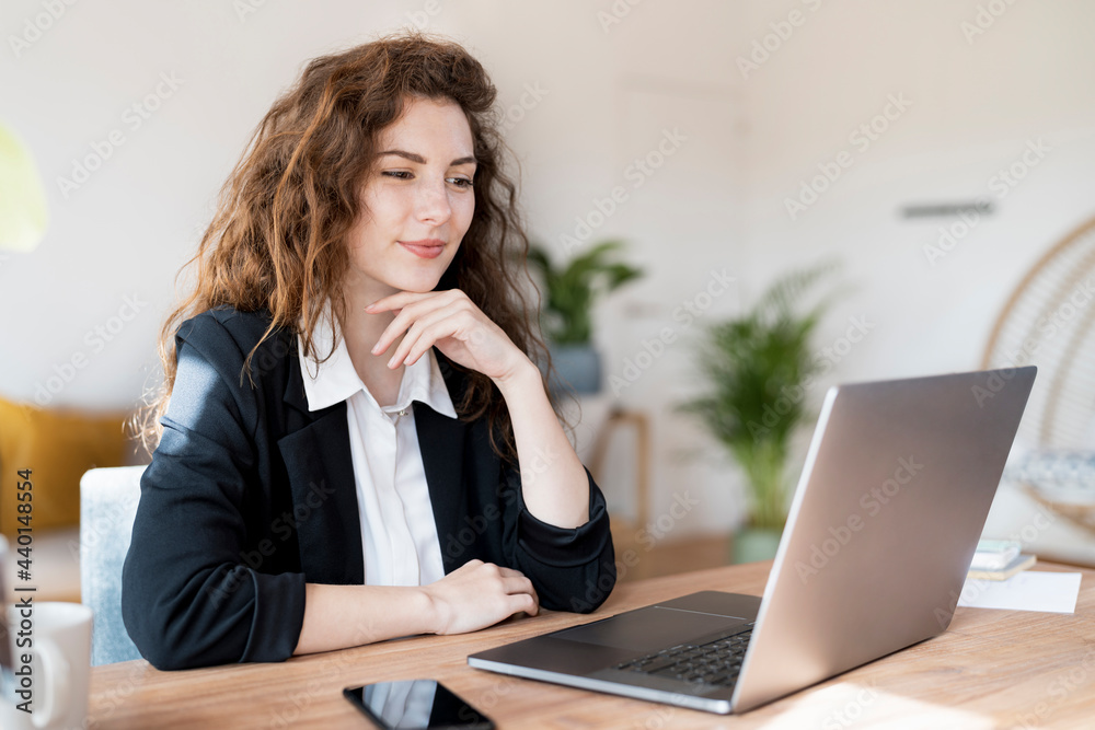 Businesswoman with hand on chin looking at laptop in home office Businesswoman with hand on chin looking at laptop in home office