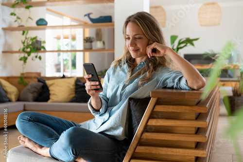 Blond hair woman text messaging through smart phone while sitting in living room photo