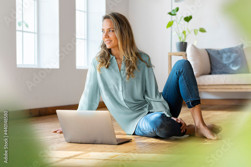 Smiling woman looking away while sitting on floor in front of laptop at home photo