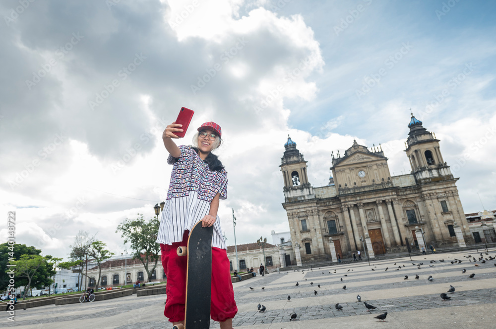 Fototapeta premium A Girl with her skateboard takes a selfie with her cell phone in front of the church.