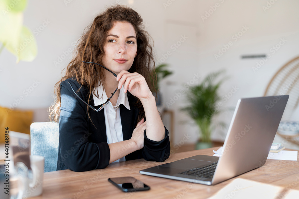 Thoughtful female entrepreneur looking away while holding eyeglasses at desk Thoughtful female entrepreneur looking away while holding eyeglasses at desk