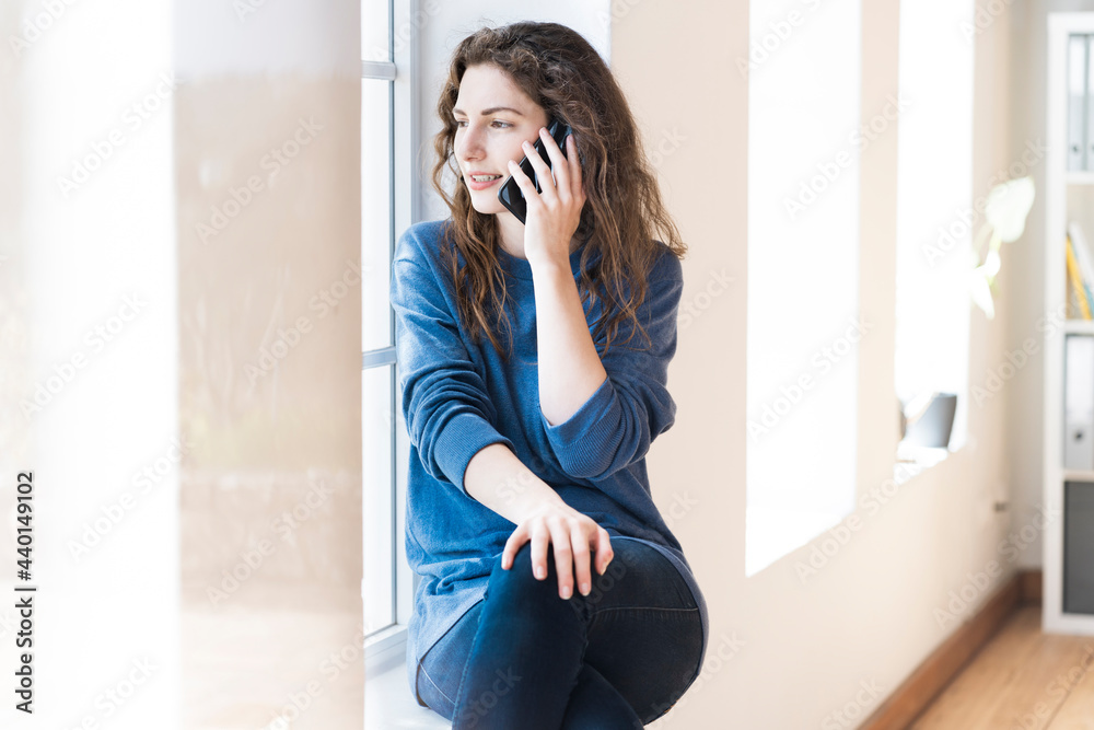 Young woman looking through window while talking on mobile phone at home Young woman looking through window while talking on mobile phone at home