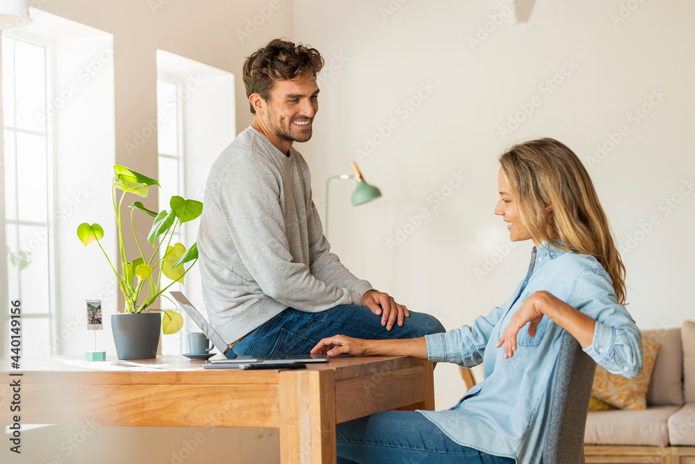 Man discussing with girlfriend looking at laptop while sitting on desk at home office Man discussing with girlfriend looking at laptop while sitting on desk at home office