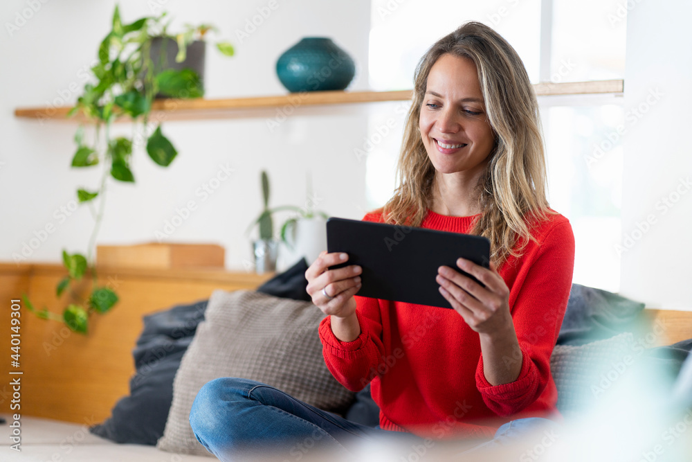 Smiling blond woman using tablet while sitting on couch in living room Smiling blond woman using tablet while sitting on couch in living room