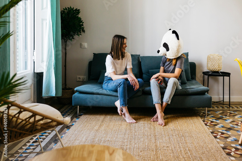 Smiling woman looking at girlfriend wearing panda mask in living room