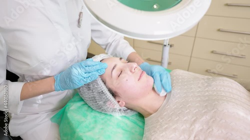 Young smiling woman enjoying beauty procedure, beautician cleaning face of female client with cleansing solution during cosmetology treatment in clinic, preparing skin before fruit acid peeling photo