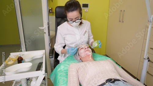 Professional beautician in mask applying cream on face skin of young woman client after fruit acid peeling, female receiving anti aging cosmetic procedure in beauty clinic. Facial rejuvenation concept photo