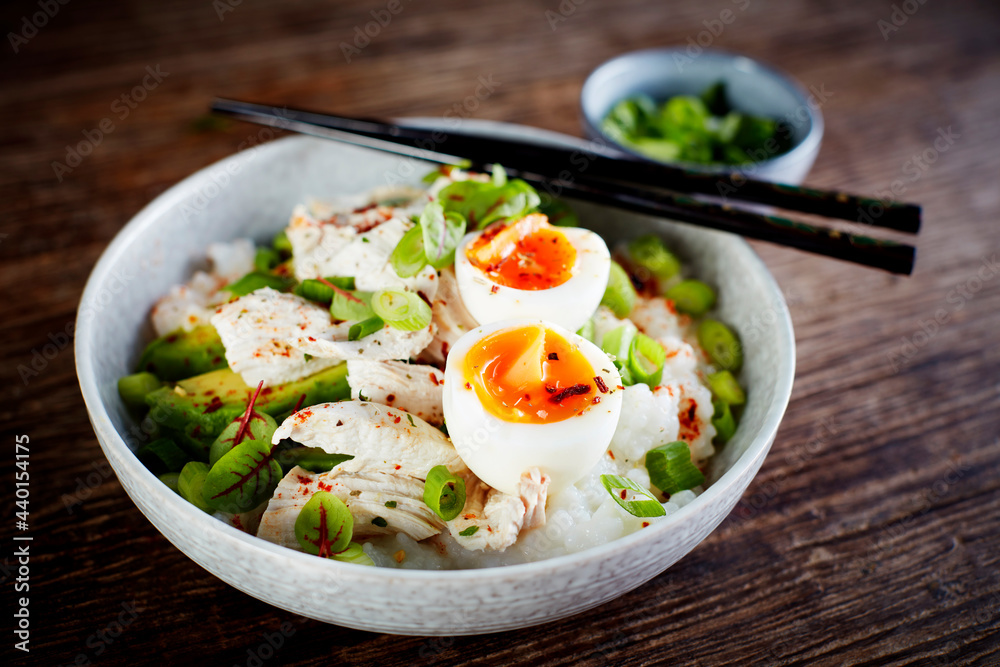 Bowl of congee with chicken breasts, boiled egg, avocado, scallions and herbs