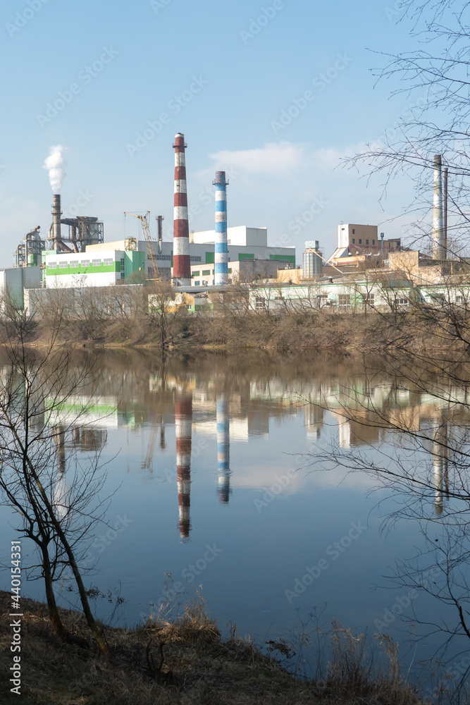 A large factory is located on the river bank. Toxic white smoke escapes from the factory's chimneys. The risk of an environmental disaster. Reflection in the water of the river of the plant