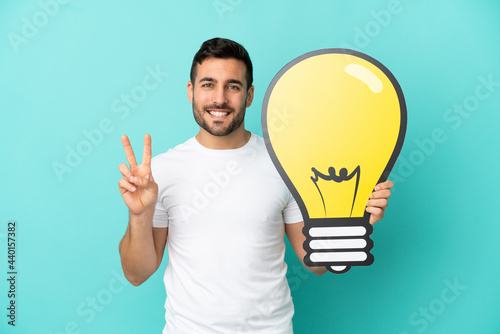 Young handsome caucasian man isolated on blue background holding a bulb icon and celebrating a victory