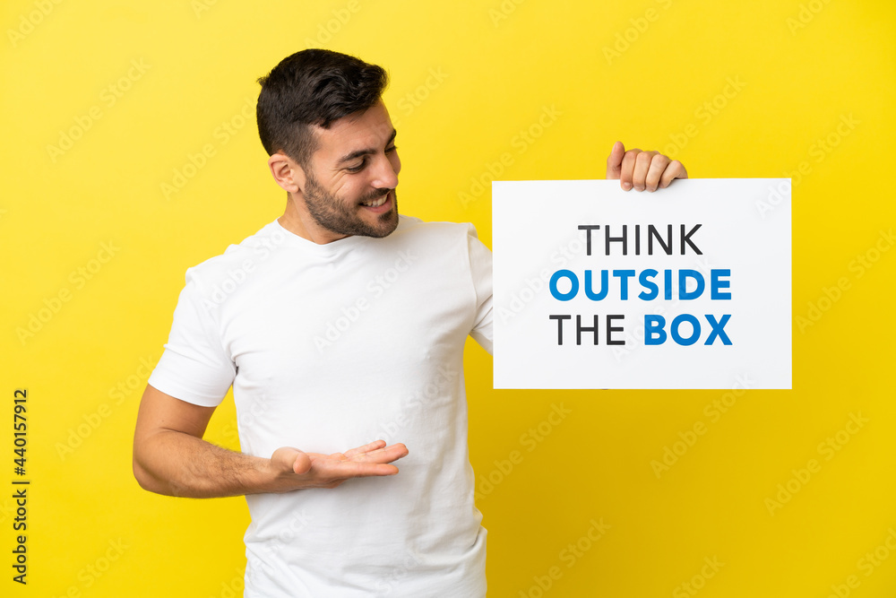 Young handsome caucasian man isolated on yellow background holding a placard with text Think Outside The Box and pointing it Young handsome caucasian man isolated on yellow background holding a placard with text Think Outside The Box and pointing it