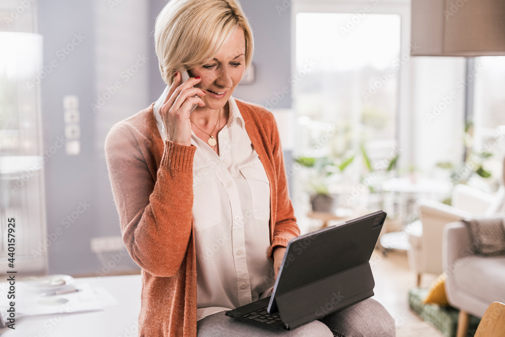 Female entrepreneur talking on smart phone while working on laptop at home Female entrepreneur talking on smart phone while working on laptop at home