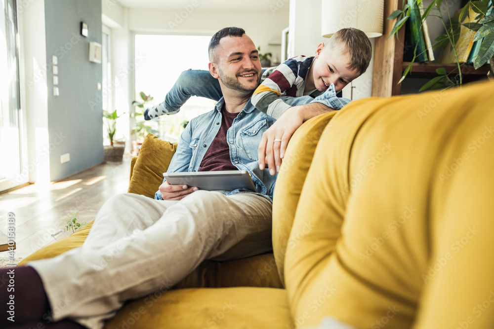 © Uwe Umstatter/Westend61 - Smiling man sitting with tablet looking at son playing in living room at home