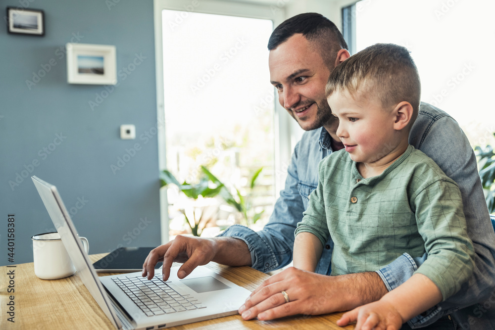 © Uwe Umstatter/Westend61 - Smiling male freelancer using laptop while sitting with son at home © Uwe Umstatter/Westend61 - Smiling male freelancer using laptop while sitting with son at home