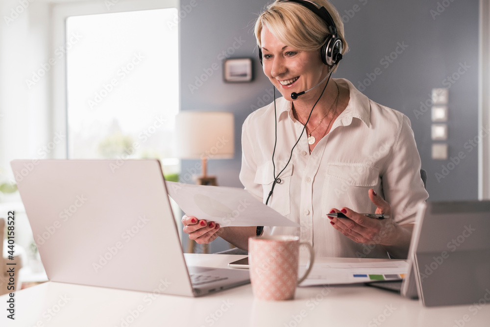 Female professional with paper talking on video call through laptop at home office Female professional with paper talking on video call through laptop at home office
