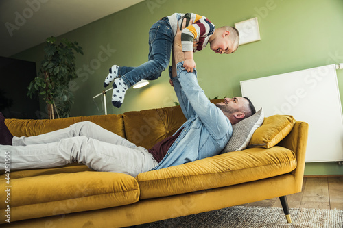 Playful father lifting up son while lying on sofa in living room at home