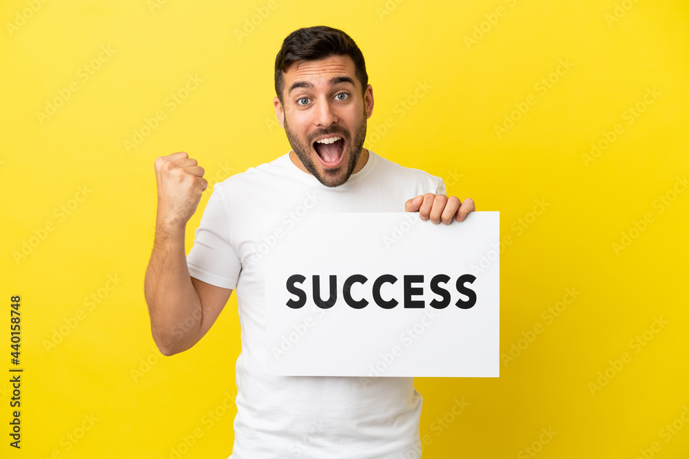 Young handsome caucasian man isolated on yellow background holding a placard with text SUCCESS and celebrating a victory Young handsome caucasian man isolated on yellow background holding a placard with text SUCCESS and celebrating a victory