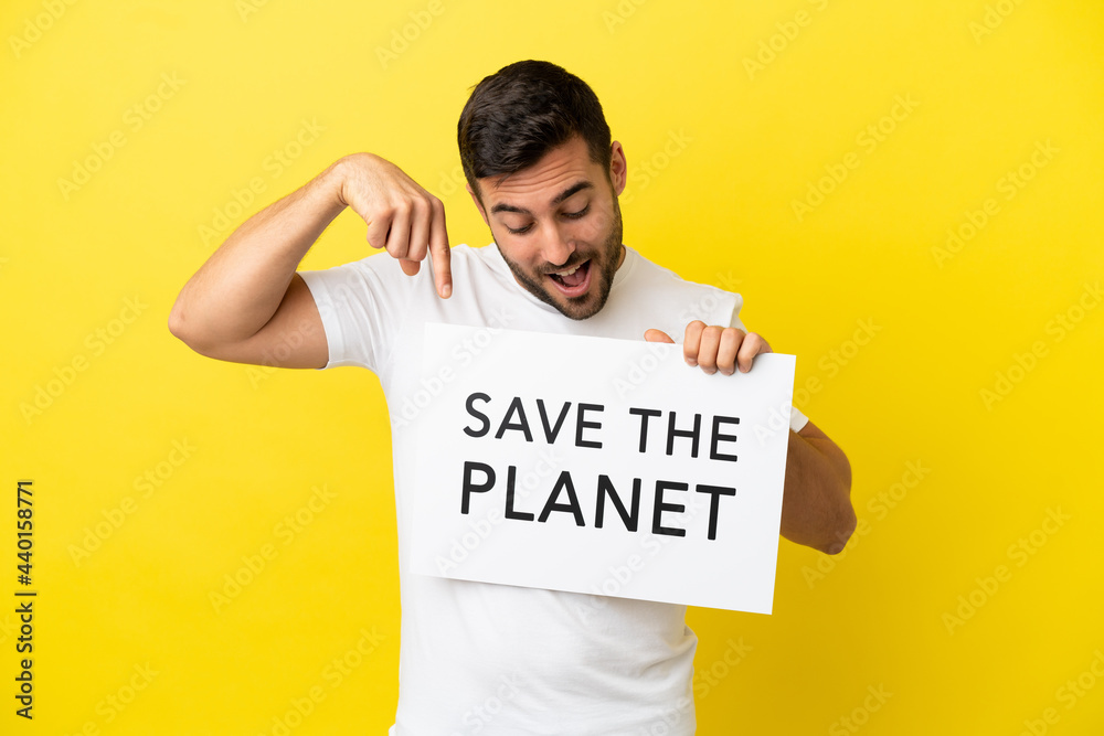 Young handsome caucasian man isolated on yellow background holding a placard with text Save the Planet and pointing it Young handsome caucasian man isolated on yellow background holding a placard with text Save the Planet and pointing it