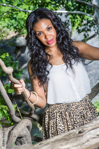 Holding on rattans in woods, a pretty black woman with long curly hair is charmingly looking at you.. photo