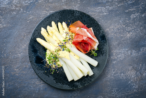 Modern style traditional steamed white asparagus with cured ham and garnished with sauce hollandaise served as top view on a Nordic design plate with copy space